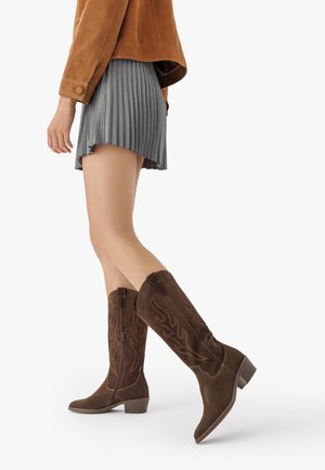 Donna che indossa una giacca in suede marrone, una gonna plissettata grigia e stivali in suede marrone alti fino al ginocchio con decorazioni a fiamma cucite, mentre cammina a metà passo.