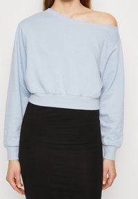 Sudadera corta azul clara con un diseño de un solo hombro, mangas largas, dobladillo acanalado y textura de algodón suave, combinada con una falda negra.