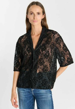 Donna che indossa un top nero in pizzo floreale con bottoni e maniche tre quarti e jeans blu su uno sfondo chiaro semplice.