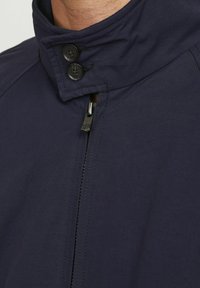 Chaqueta de marina con un alto cuello, botones visibles y cierre frontal con cremallera. Está hecha de un tejido duradero con una textura suave y un detalle mínimo.