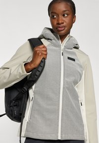 Veste légère avec un design rayé gris et blanc, dotée d'une capuche, fabriquée à partir d'un matériau doux, complétée par une fermeture éclair noire et des poches.
