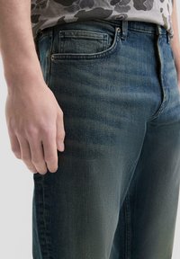 Jeans in denim blu scuro con una finitura leggermente sbiadita, dotati di cinque tasche, passanti per la cintura e un design slim fit. Texture liscia.