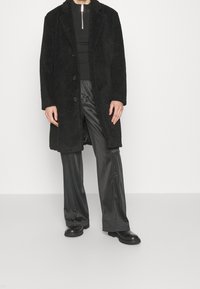 Sort faux fur frakke med klapkrave, front knapper, parret med skinnende sorte bukser og robuste sorte støvler. Ensfarvet baggrund.
