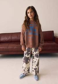 Sweatshirt marron oversize avec texte bleu, pantalon large à motifs blanc et marron, et baskets bleu clair, debout sur un sol beige.