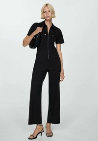 Sort jumpsuit med korte ærmer, lynlås foran og lommeaccenter; har en tætsiddende bodice og vide bukser, parret med sorte strophæle.