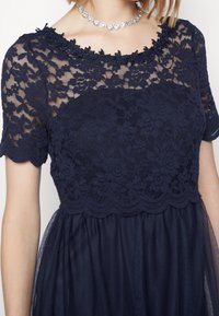 Mörkblå spetsklänning med blommönster, vågig halsringning och korta ärmar, samt en svävande genomskinlig kjol. Utsmyckad med ett silverhalsband.