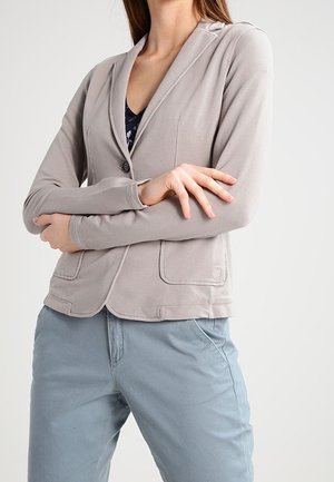 Vrouw met gekruiste armen die een lichtgrijs colbert draagt over een donker patroon blouse en lichtblauwe broek, tegen een eenvoudige witte achtergrond.