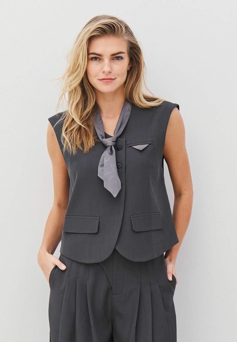 Blazer gri fără mâneci, cu un design scurtat, două buzunare frontale și o eșarfă gri asortată legată la gât, cu o textură netedă.