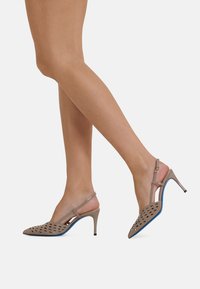 Beige slingback heels with a pointed toe, featuring cut-out patterns, a blue outsole, and a slim heel. Straps provide secure fit.