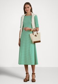 Zöld-fehér csíkos midi ruhát viselő nő, vállán fehér kábelmintás pulóvert lógatva, bézs színű bevásárlótáskát hord, barna ék sarukban.