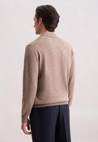 Mann mit dunklen Haaren, der einen beigen Strickpullover und dunkelblauen Hosen trägt, mit dem Rücken zu einem schlichten hellen Hintergrund.