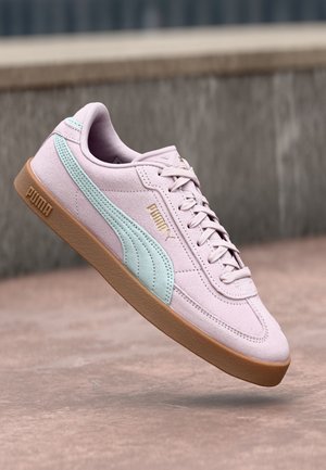 Hellrosa Wildleder-Puma-Sneaker mit mintgrünem Seitenstreifen, goldenem Logo, Gummisohle und Schnürsenkeln, in der Luft vor Betonhintergrund gezeigt.