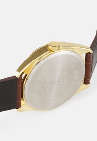 Gold watch case with a stainless steel back, featuring a brown leather strap. The crown is located at the 3 o’clock position.