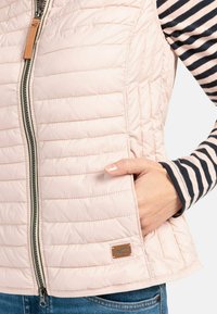 Lichtroze gewatteerde vest met een rits aan de voorkant, zakken aan de zijkant en een bruin leren label. Gestreept overhemd met lange mouwen zichtbaar eronder.