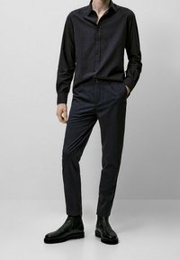 Dunkel gestreiftes Hemd mit Knöpfen, kombiniert mit dunkel geschnittenen, schmalen Hosen und schwarzen Stiefeletten. Glatter Stoff mit dezenten Mustern und strukturierter Passform.