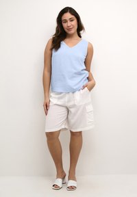 Haut sans manches bleu clair avec un col en V, associé à un short blanc arrivant aux genoux avec des poches latérales. Modèle portant des sandales blanches à glisser.