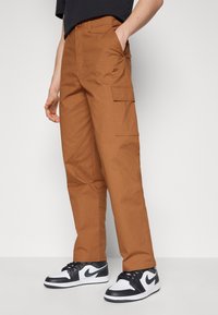 Bruna cargobyxor med en texturerad tyg, med en liten Nike-logotyp och sidofickor, ihop med svarta och vita Nike-sneakers.