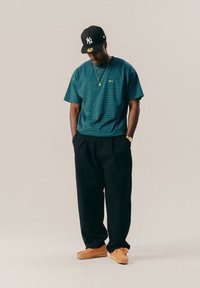 Férfi fekete New York Yankees sapkában, kék csíkozott ingben, fekete nadrágban, narancssárga cipőben és arany kiegészítőkkel áll, keze a zsebében, lefelé nézve.