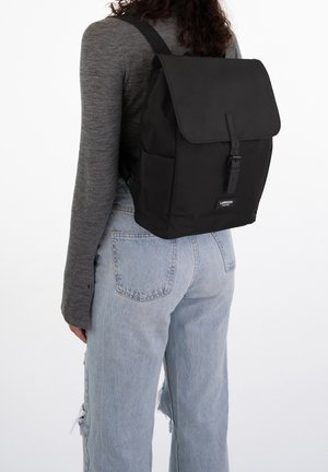 Henkilö, jolla on päällä vaaleansiniset revityt farkut ja harmaa pitkähihainen paita, kantaa mustaa reppu, jossa on läppä ja solkilukko.