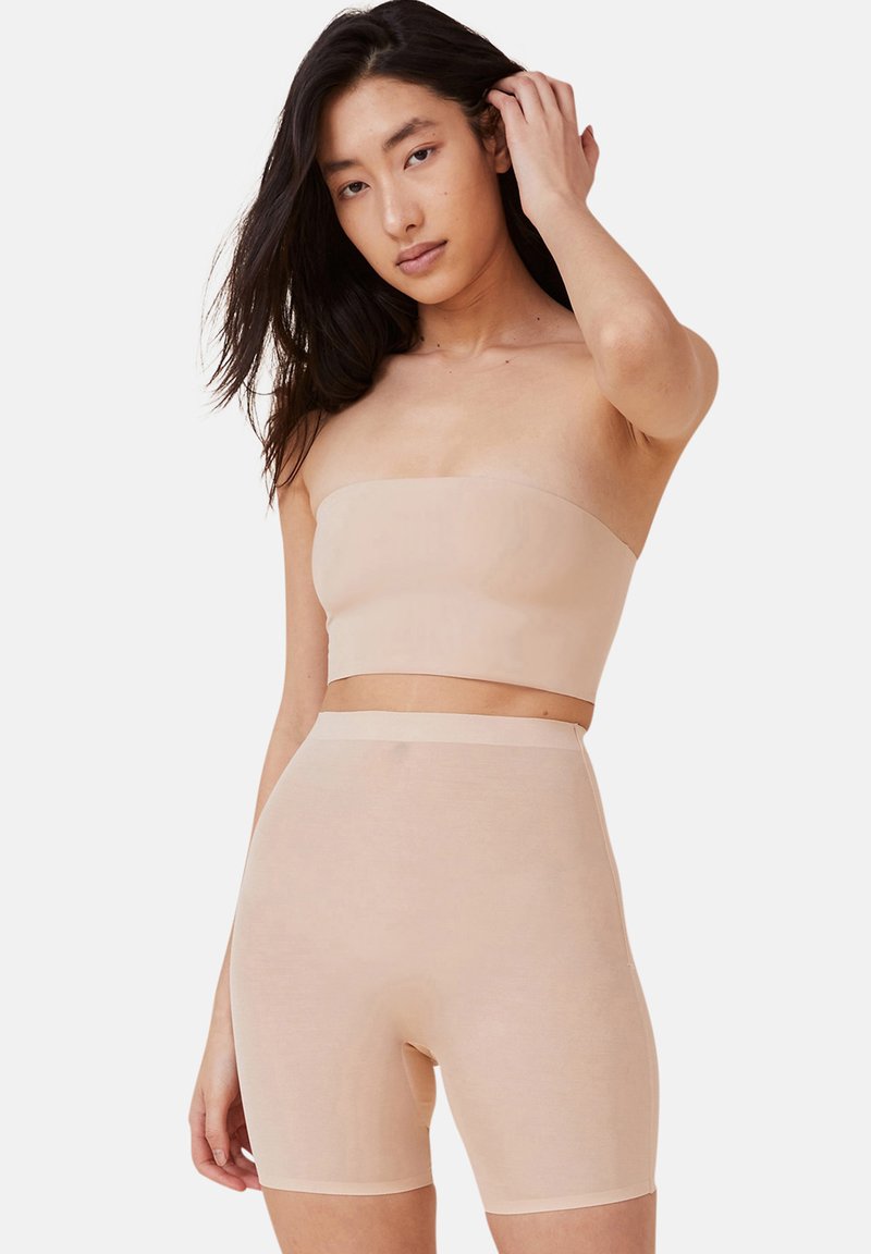 Kvinna med långt mörkt hår som bär en hudfärgad strapless topp och högmidjade cykelshorts, poserande med en hand som rör vid sitt hår.