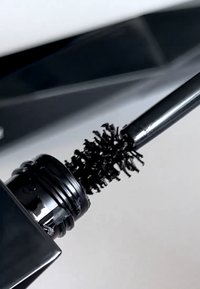 Czarna szczoteczka do tuszu do rzęs z skręconą, puszystą końcówką, wychodząca z eleganckiej czarnej tubki o błyszczącym wykończeniu i szczegółowych pierścieniowych ozdobach.