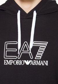 Svart hoodie med vita snören. Har en stor vit "EA7" logo och texten "EMPORIO ARMANI" på framsidan. Mjukt tyg, avslappnad design.