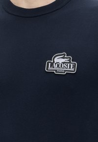 Gros plan sur un tissu bleu marine avec un patch logo Lacoste Paris présentant un crocodile blanc sur la zone de la poitrine d’un vêtement.