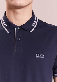 Polo-shirt marine en coton, avec un col classique à rayures grises et blanches, deux boutons, et un logo brodé blanc.