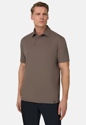 Mann trägt ein kurzärmeliges braunes Polohemd und schwarze Hose, steht mit neutralem Gesichtsausdruck vor einem schlichten weißen Hintergrund.