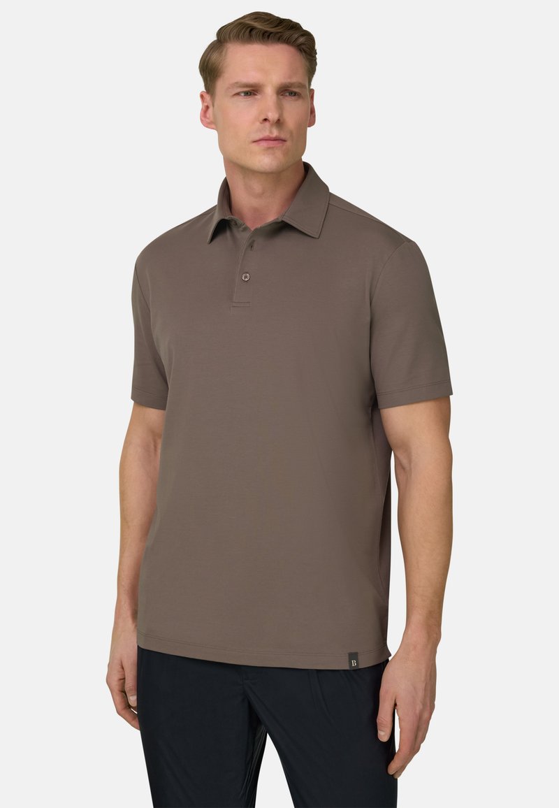 Homme portant un polo marron à manches courtes et un pantalon noir, debout avec une expression neutre sur un fond blanc uni.