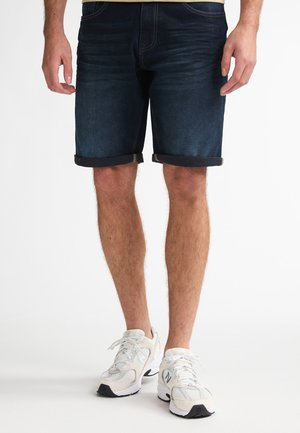 Donkerblauwe denim shorts met een slanke pasvorm, opgerolde zoom en subtiele vlekkering. Gecombineerd met witte sportschoenen met een zwart accent.