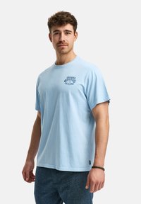 Jeune homme avec des cheveux bouclés portant un t-shirt bleu clair avec le texte "Votre énergie est guérissante", se tenant devant un fond uni clair.