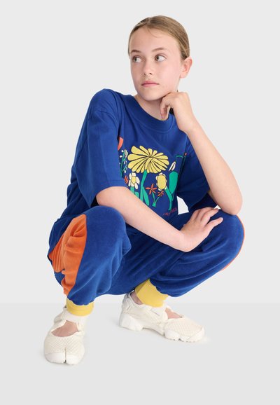 Junge Person in Hockposition, trägt ein blaues Hemd mit bunten Blumen, passende blaue Hose mit orangefarbenen Flicken, gelbe Socken und weiße Schuhe.