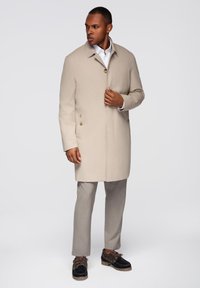 Cappotto beige con colletto e due tasche frontali, caratterizzato da una chiusura con bottoni e una consistenza liscia, abbinato a pantaloni grigi e scarpe nere.