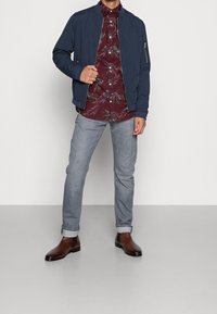 Granatowa kurtka typu bomber, czerwona koszula w kwiaty zapinana na guziki, jasnoszare dżinsy, brązowe skórzane buty typu chelsea. Gładkie materiały, luźny krój, podwinięte nogawki dżinsów.