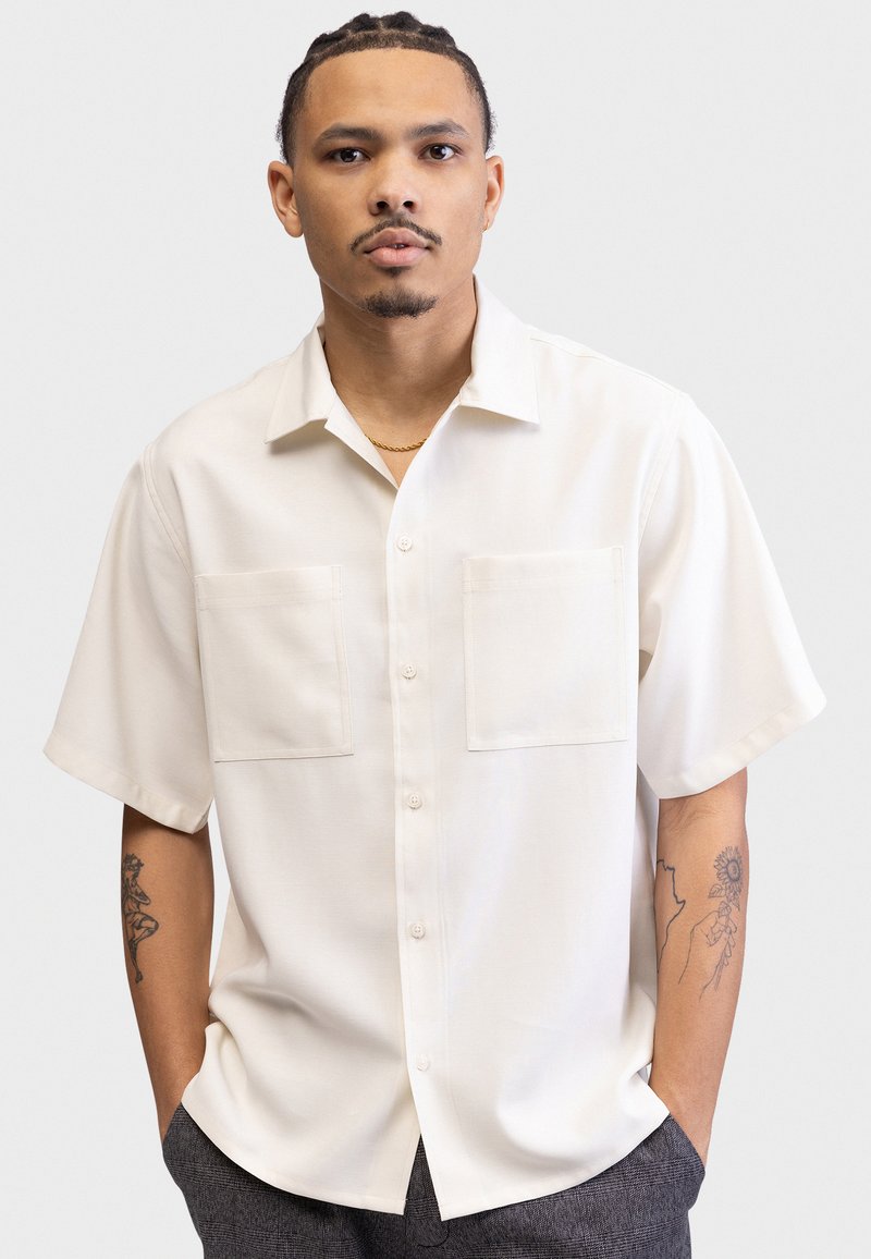 Light cream short-sleeve button-up shirt with two chest pockets and a soft, smooth texture. Features a classic collar and a relaxed fit.