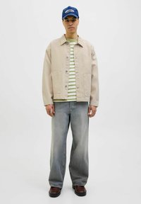 Beige jacket, striped green and white shirt, light blue jeans, brown shoes, and a blue cap with white text. Model stands against a plain background.