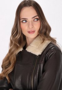 Giacca in pelle marrone con colletto in pelliccia sintetica beige, dotata di zip frontale e spalle leggermente strutturate. Texture liscia e design casual.