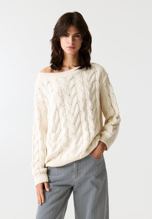 Maglione a trecce color crema con design senza spalle e maniche lunghe, abbinato a pantaloni a gamba larga a righe. Texture naturale e calore.