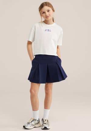 Jeune fille aux cheveux blonds portant un t-shirt blanc court avec texte, une jupe plissée bleu marine, des chaussettes blanches et des baskets argentées, debout les mains dans les poches.