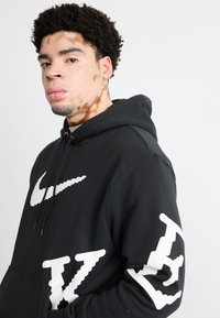 Μαύρο φούτερ από μείγμα βαμβακιού, με μεγάλο λευκό λογότυπο Nike στο στήθος και λευκή γραφή στο μανίκι. Υφή με ανάγλυφη επιφάνεια.