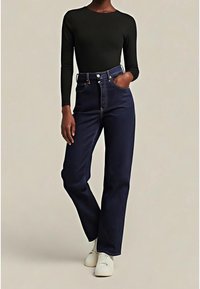Personne portant un haut noir à manches longues, un jean bleu foncé taille haute, une main dans la poche, et des baskets blanches, debout devant un fond uni.