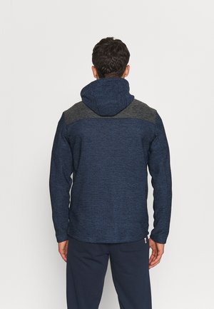 Uomo che indossa una giacca con cappuccio blu scuro con pannelli grigi sulle spalle, in piedi con le spalle rivolte verso una sfondo chiaro uniforme.