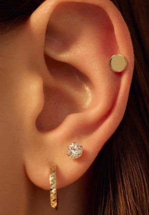 Goldene Creolenohrringe mit strukturiertem Design und ein mit Kristallen besetzter Steckerohrringe im durchstochenen Ohr, die den Hautton und die Ohrkonturen präsentieren.