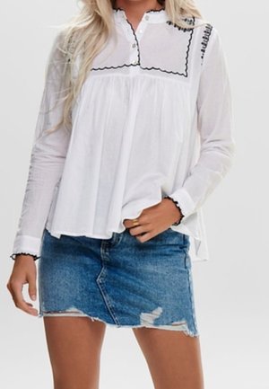 Blusa bianca con un design arricciato e bordi neri scallop, abbinata a una gonna di jeans strappata. Il tessuto è leggero e informale.