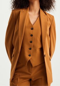 Beige tre-delars kostym med en skräddarsydd blazer, väst med fem svarta knappar och matchande byxor. Slät struktur, figursydd design.