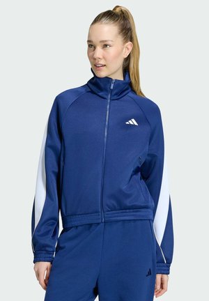 Mladá žena se světlým culíkem, která má na sobě modro-bílou sportovní bundu na zip a odpovídající kalhoty, a dívá se mírně doprava.