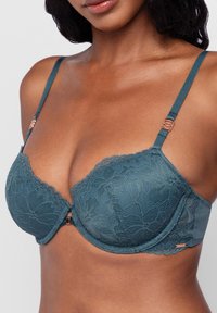 Woman wearing teal lace push-up bra with floral pattern and rose gold accents, showing upper torso and neckline on a light background.