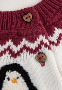 Tissu tricoté bordeaux et blanc avec motif en zigzag, boutons en bois en forme de cœur, et partie d’un motif de pingouin visible.