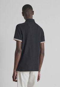 Hombre de pie de espaldas, vistiendo una camiseta tipo polo de manga corta negra con puños a rayas y pantalones blancos, frente a un fondo liso.